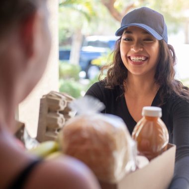 A woman wearing a cap deliveries a bag of groceries to a woman at the front door.