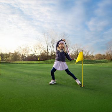 A young girl stands on a golf course and swings a golf club.
