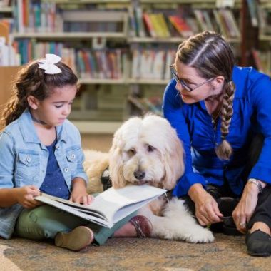 A young girl and an adult librarian woman, both dressed in blue, read a book to a dog with long white hair.
