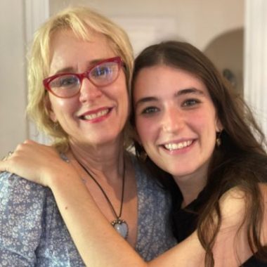 Mother with blond hair embraces adult daughter with dark brown hair.