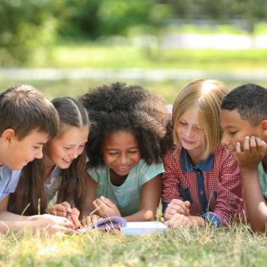 Five young children lounge on grass around a book that they are reading.