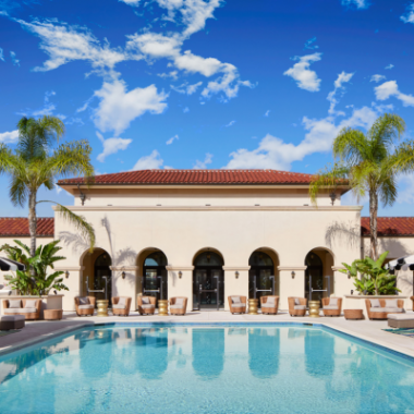 A cream-colored hotel stands behind its outdoor pool surrounded by lounge chairs. Palm trees flank the hotel.