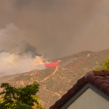 Land in the background burns in a wildfire. In the foregound is the frame of a house with Spanish tile.